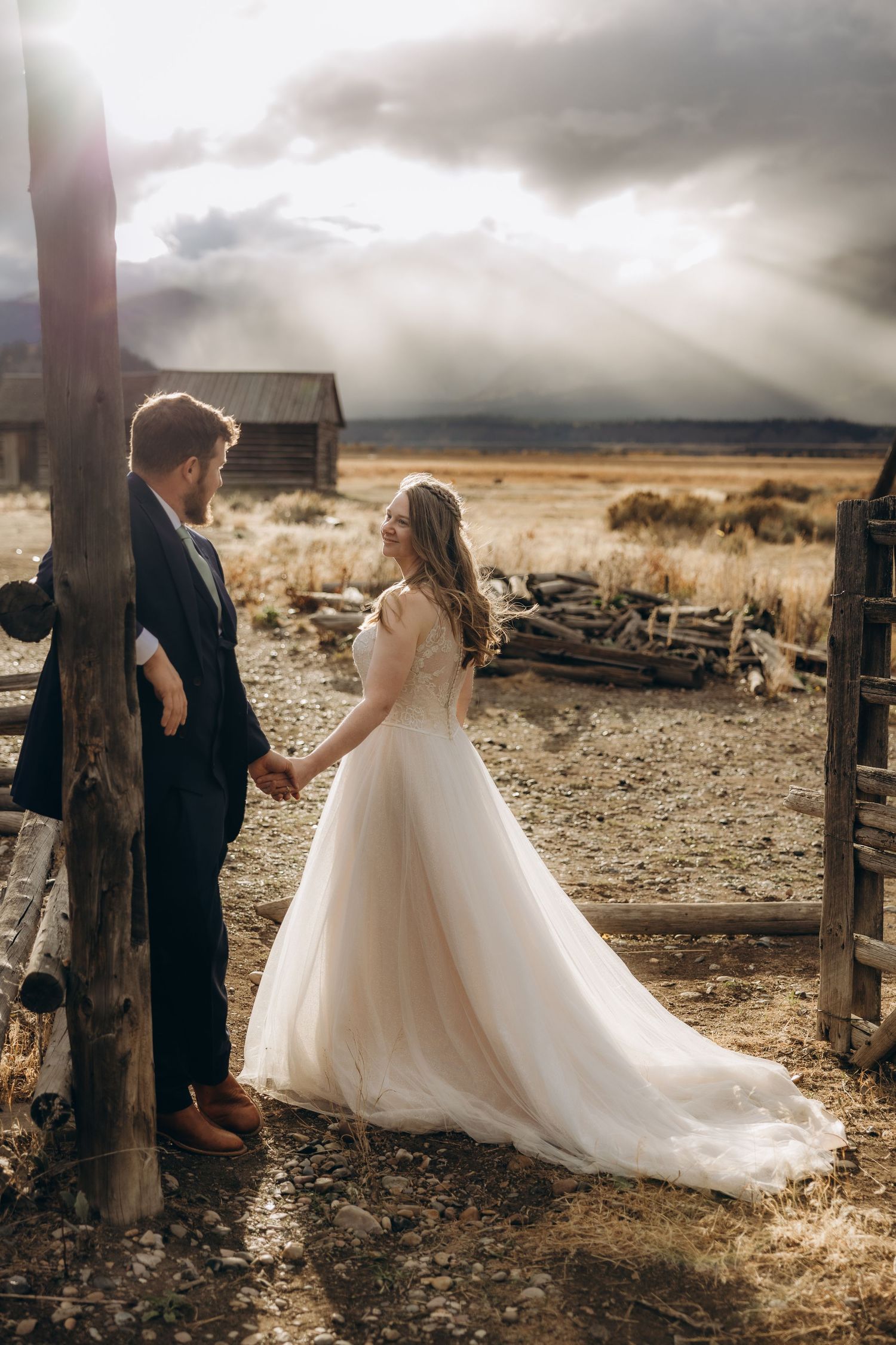 A romantic wedding couple stands between rustic wooden fence posts against a dramatic stormy sky in a desert landscape.
