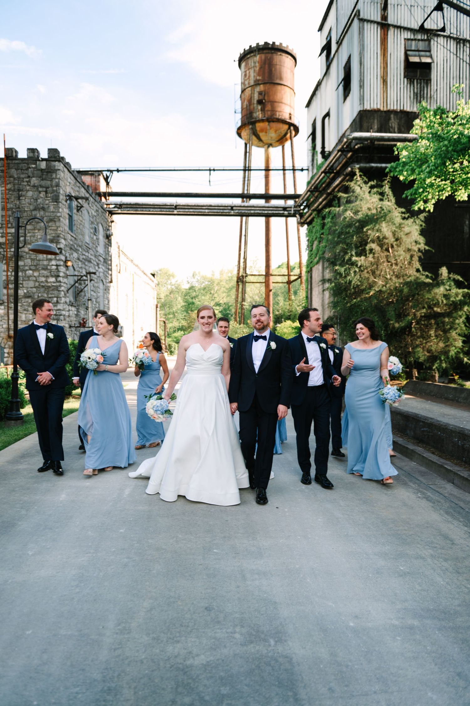 Wedding party in formal attire walking and laughing together on an industrial rooftop with rustic water tower in background.