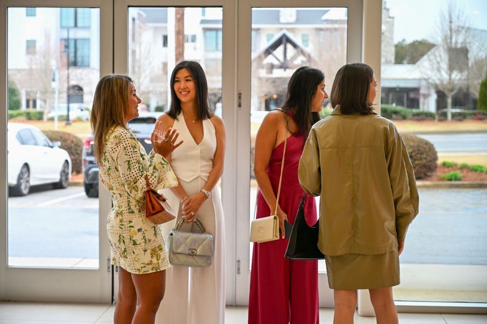 A group of women in casual dresses stand chatting near glass doors in a bright indoor space.
