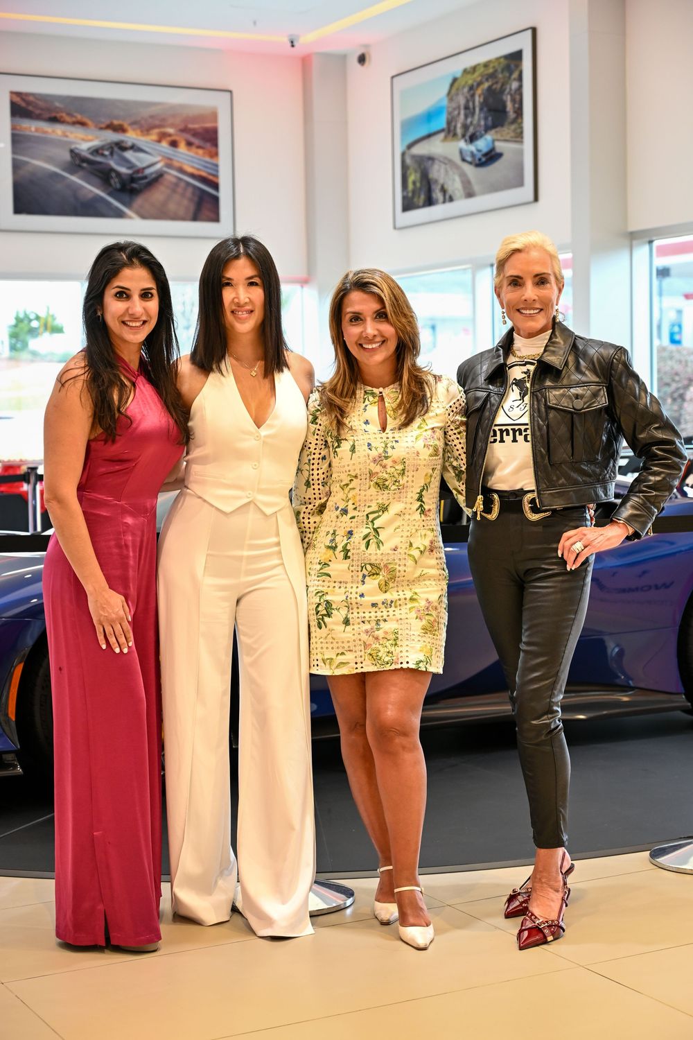 A group of women pose together in stylish outfits including a burgundy dress, white pantsuit, and leather jacket at an event.