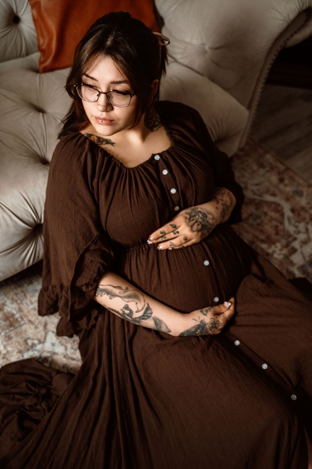 Person wearing a brown dress with long sleeves sits on a light colored couch in moody indoor lighting.