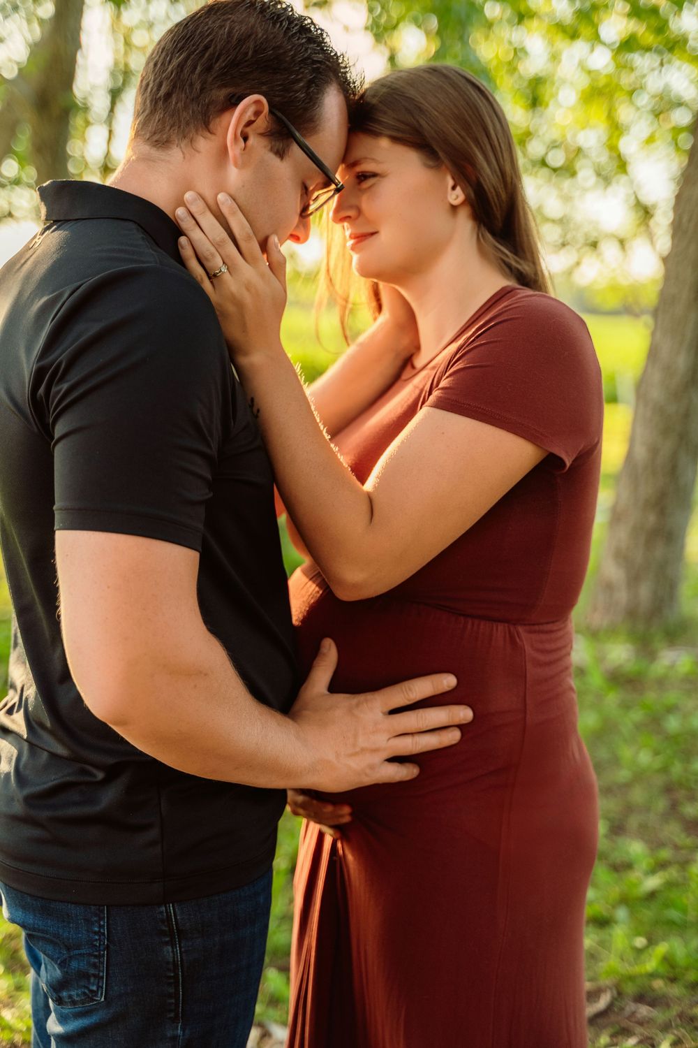 A couple shares a tender moment outdoors, with the woman in a burgundy dress and man in black embracing at golden hour.