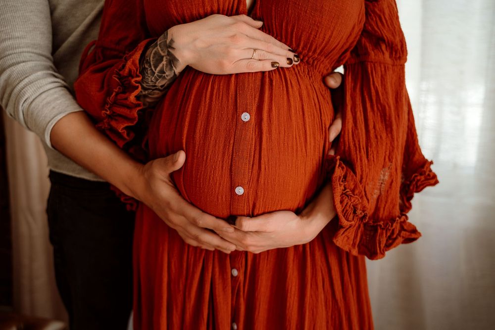 Pregnant woman in rust red dress cradling baby bump with partner's hands around her belly.