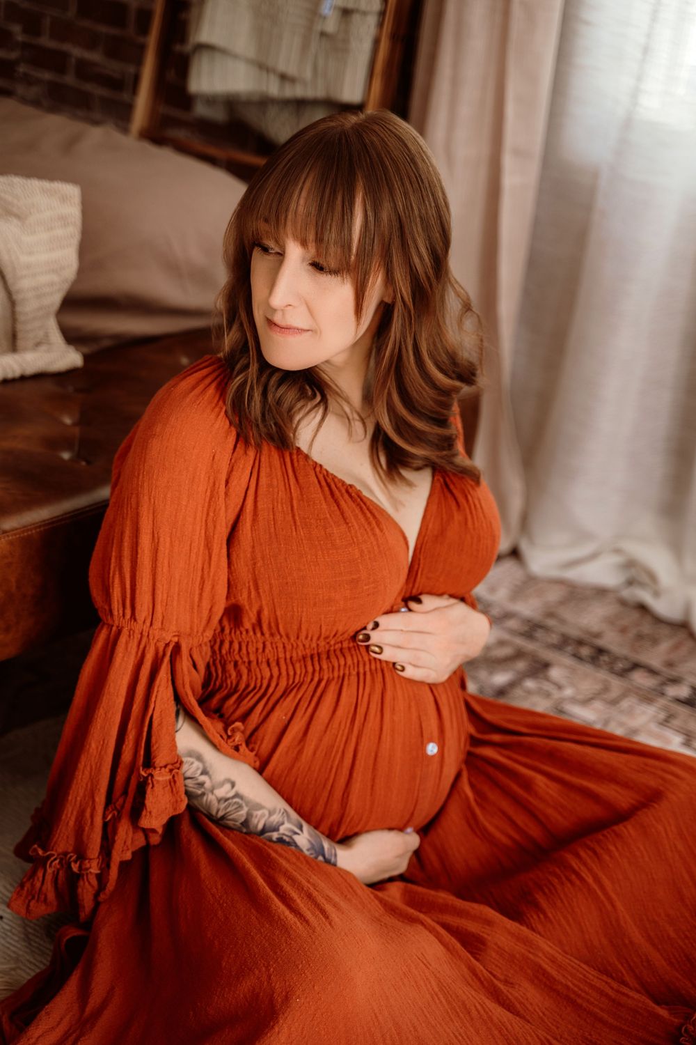 Pregnant woman in rust orange boho dress cradling baby bump, seated gracefully in a warm, softly lit maternity photo session.