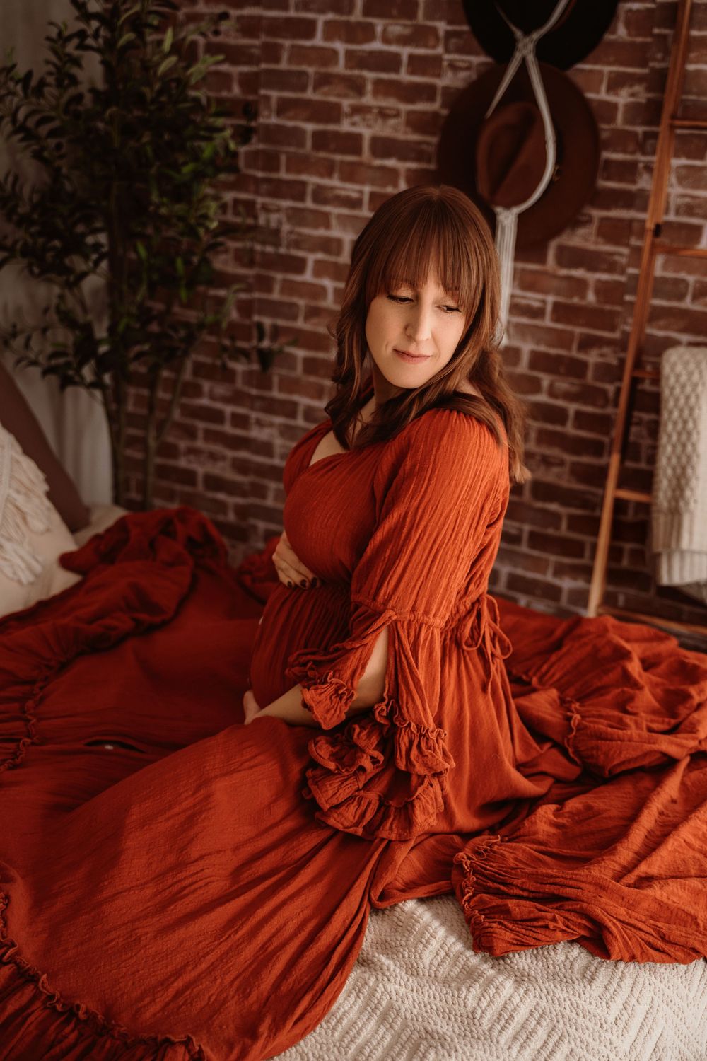 Pregnant woman in rust orange boho dress sitting on bed with brick wall backdrop.