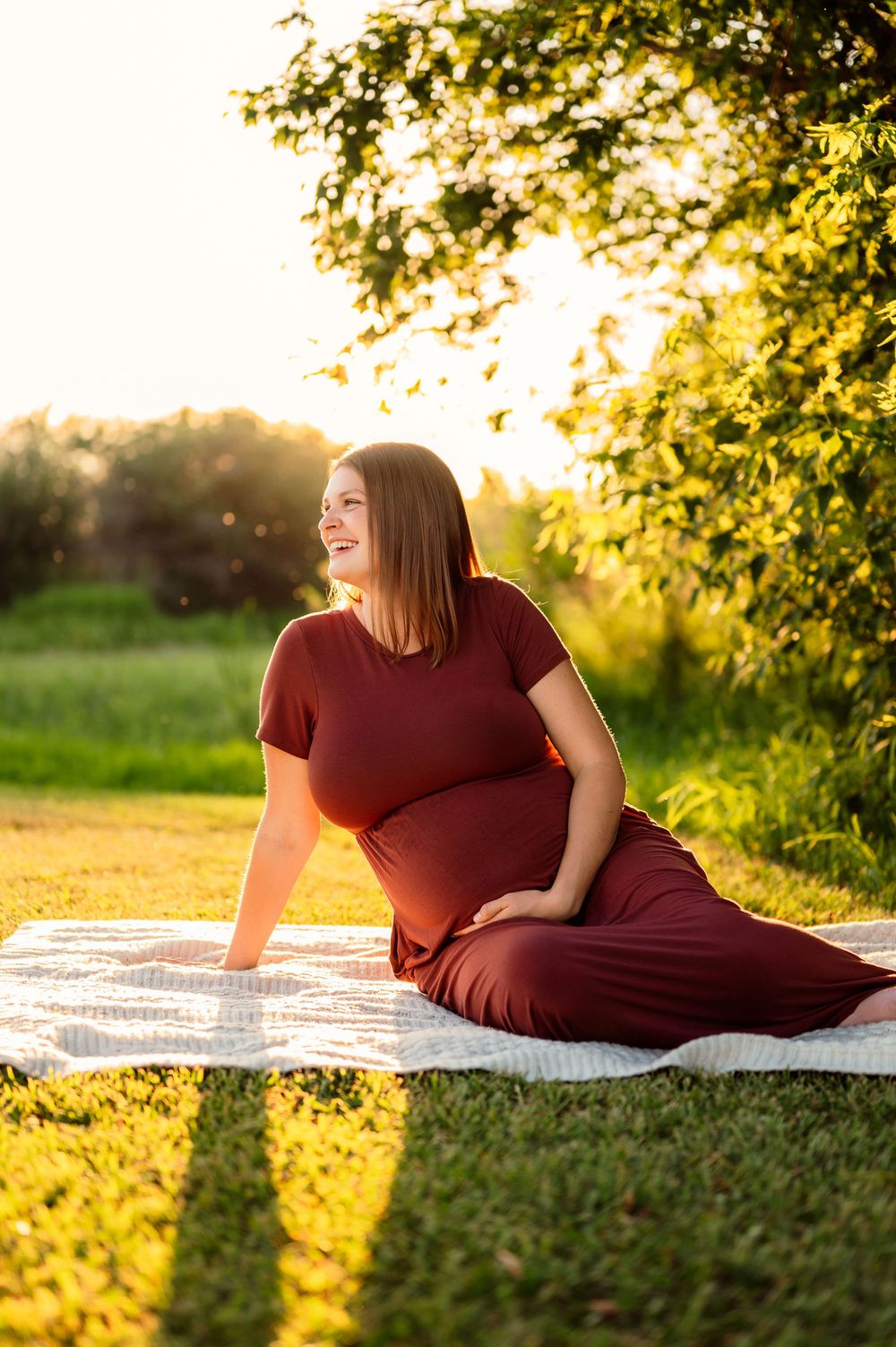 Pregnant woman in burgundy dress sitting on blanket outdoors, smiling in golden sunset light with green trees behind her.