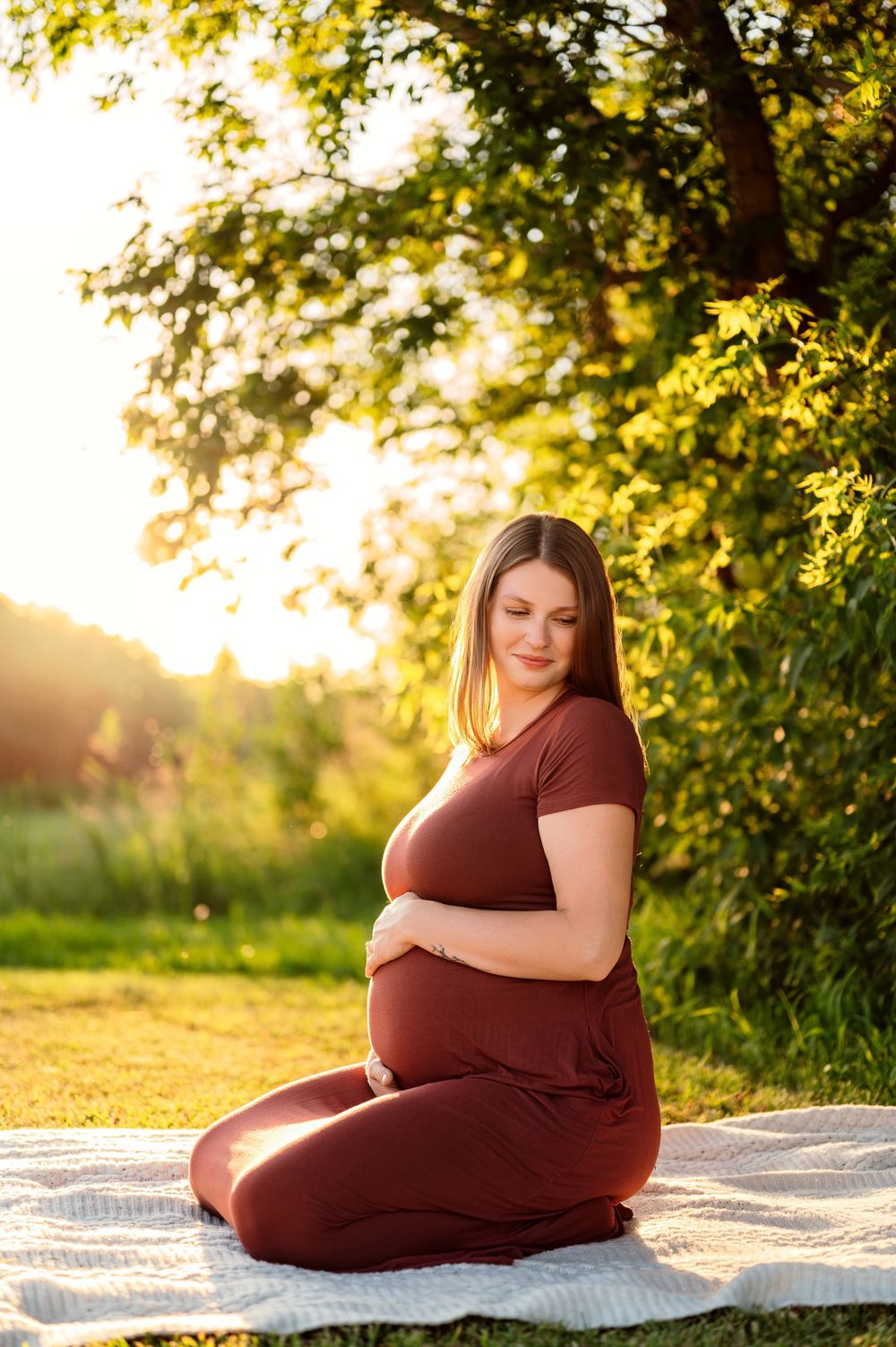 Pregnant woman in burgundy outfit sitting on blanket outdoors, cradling belly in golden sunset light.