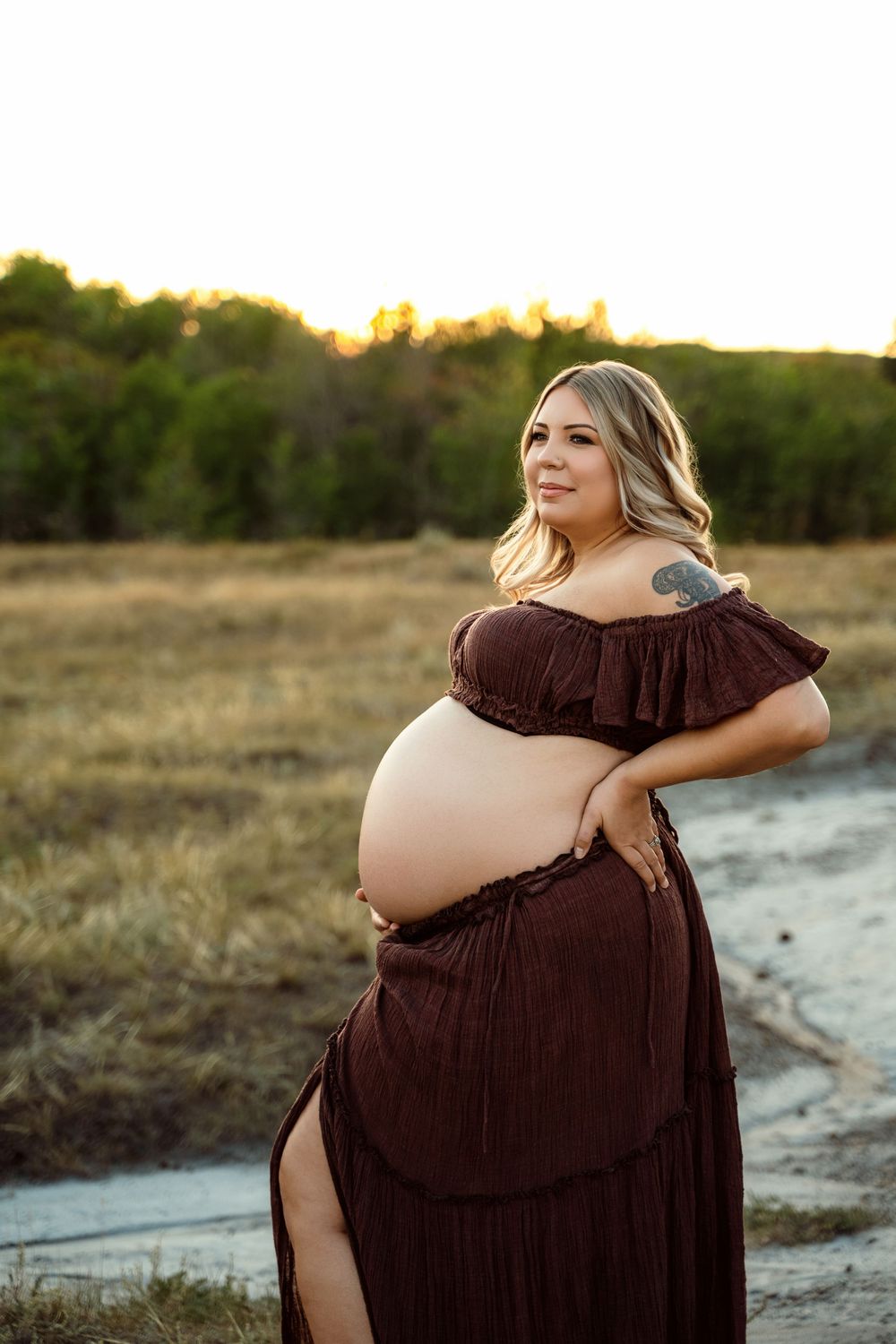 A person in a brown gown poses during a maternity photoshoot in a rustic outdoor setting at golden hour.