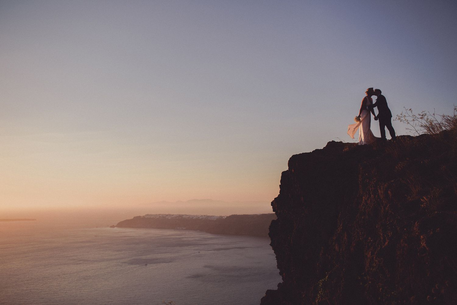 bride and groom kissing on top of cliff in santorini, greece