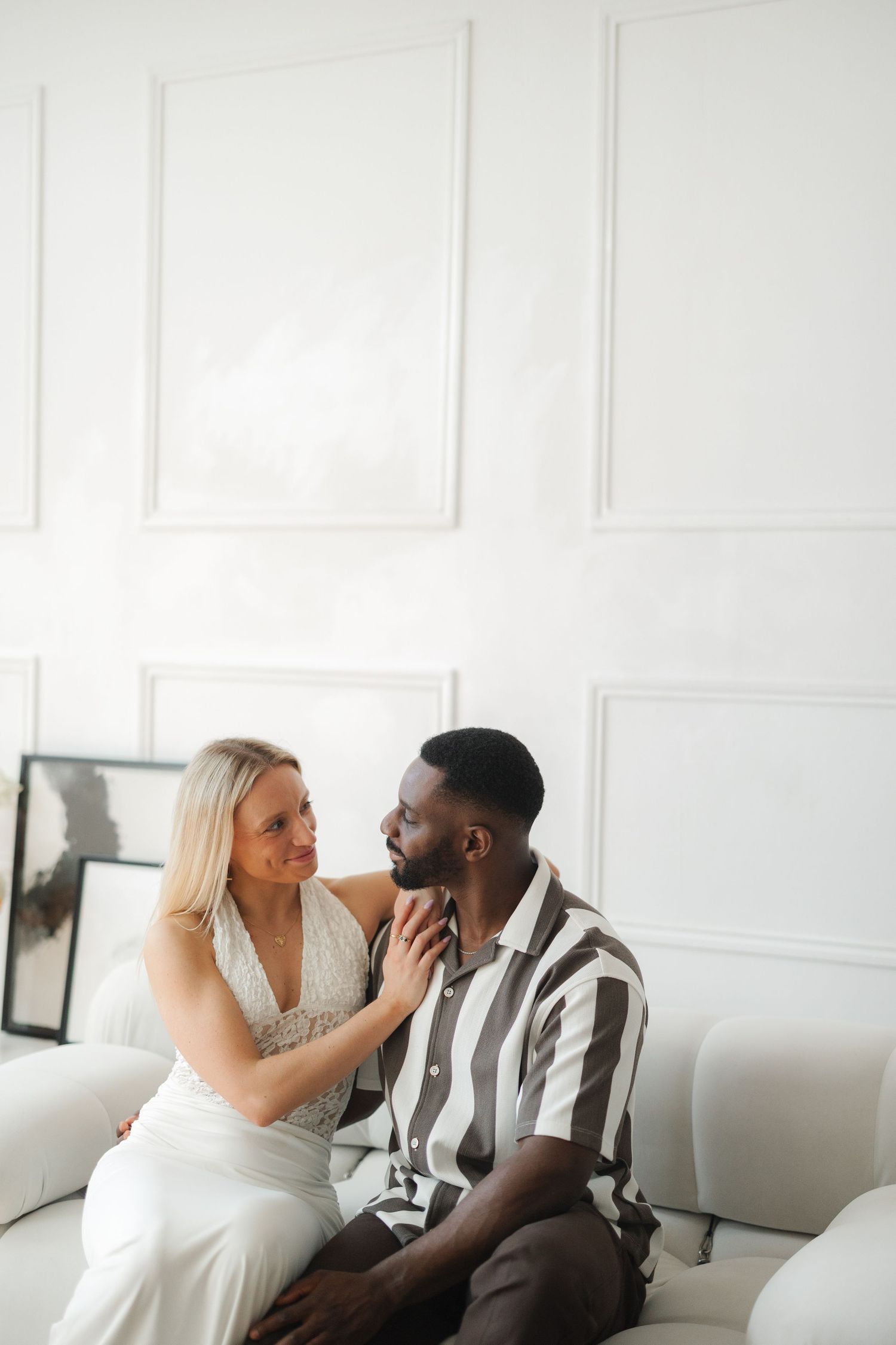 An interracial couple sits on a white sofa, sharing a tender moment in a bright, minimalist living room.