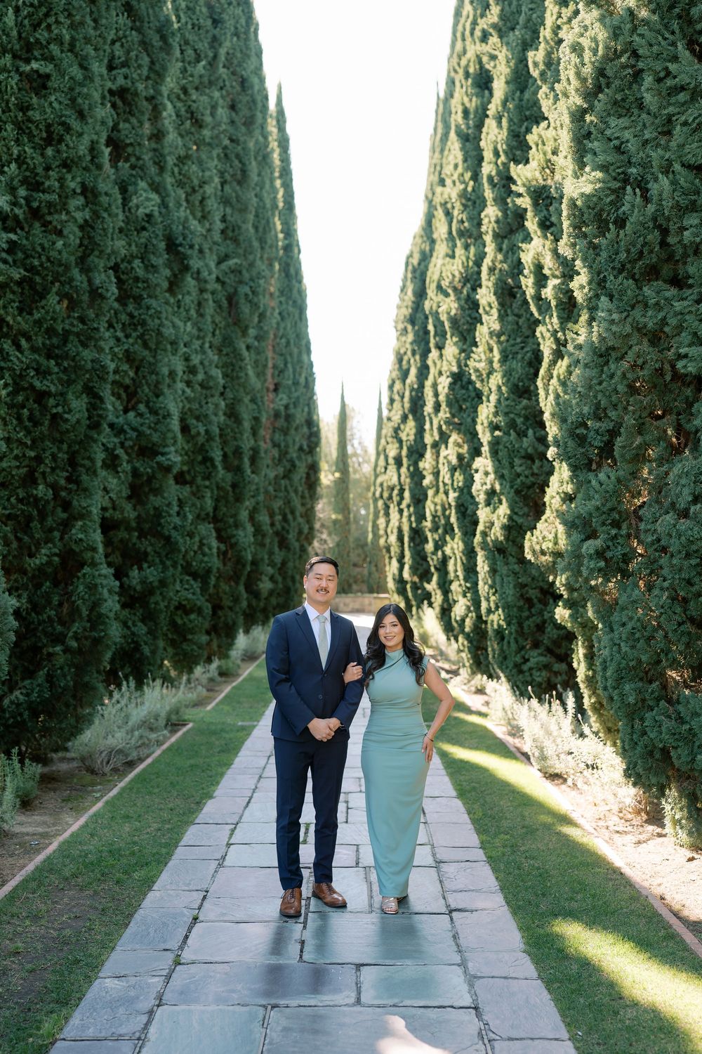 A couple walks down a scenic cypress tree-lined pathway in a garden setting during golden hour lighting.