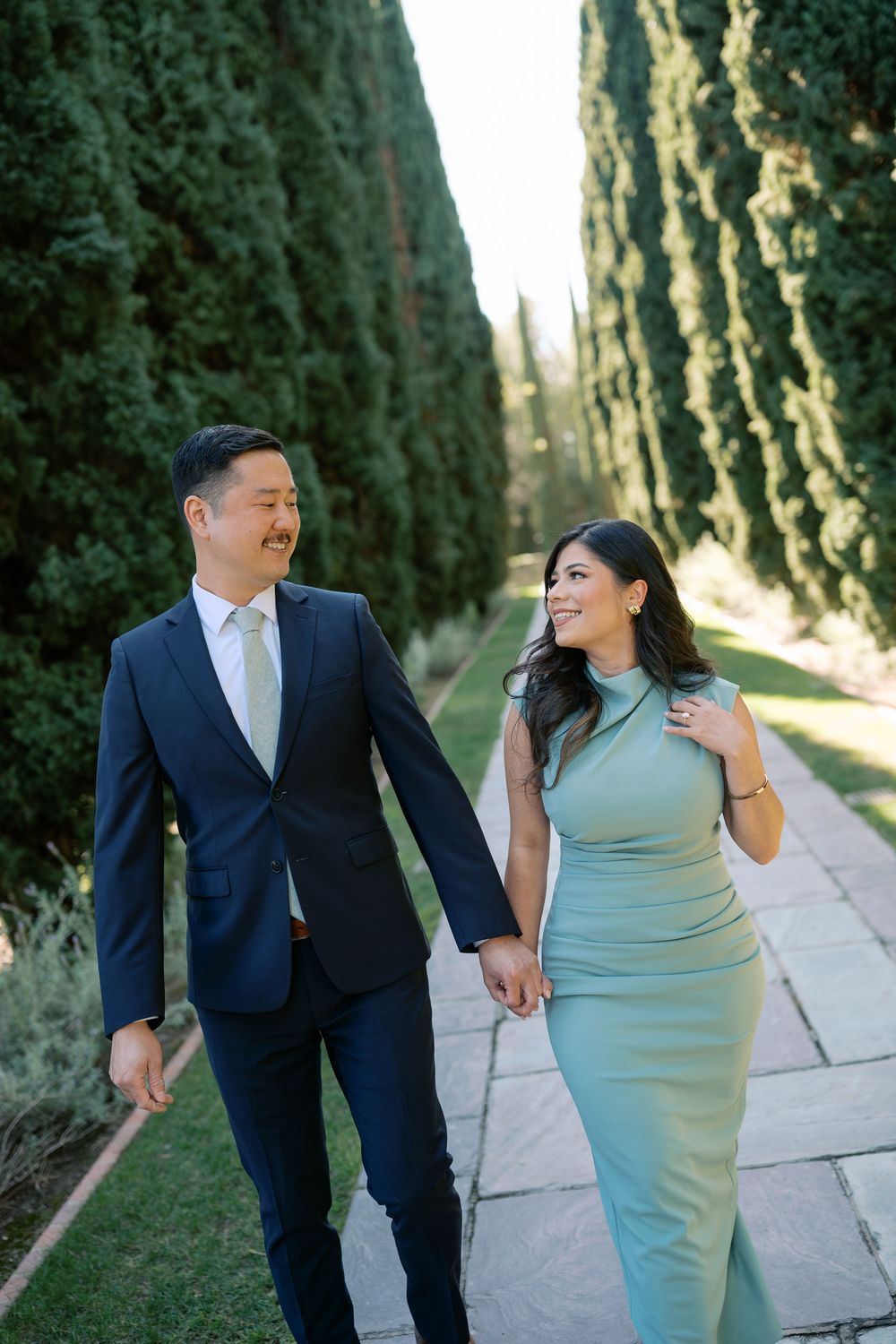 A couple in formal attire walks together and shares laughs along a tree-lined pathway during a photo session.