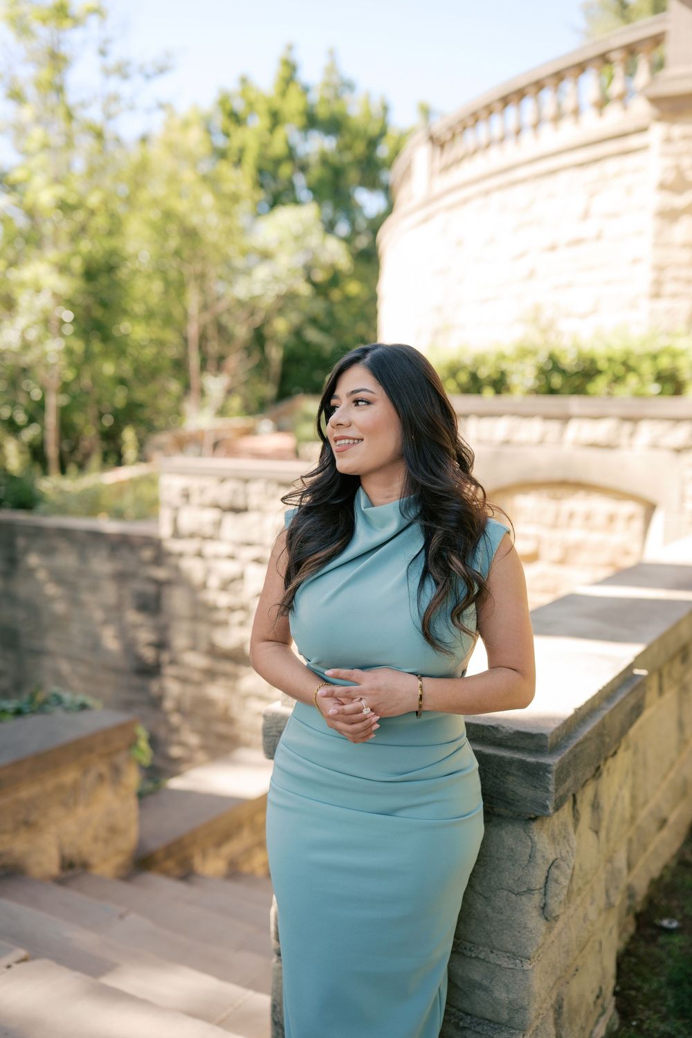 A woman in a turquoise dress poses gracefully against a stone wall backdrop in an outdoor garden setting.