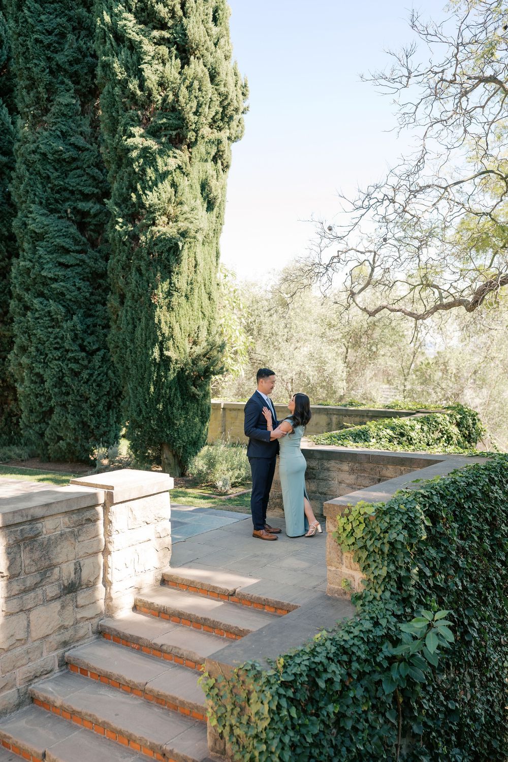 A romantic proposal moment unfolds on stone garden steps surrounded by lush cypress trees and ivy-covered walls.