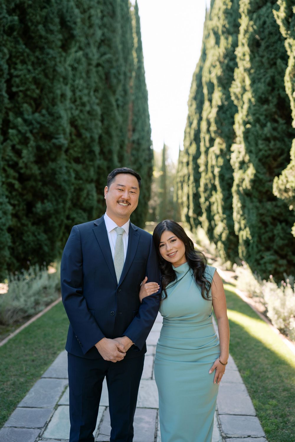 A couple shares joyful moments while walking along a cypress tree-lined path in formal attire.