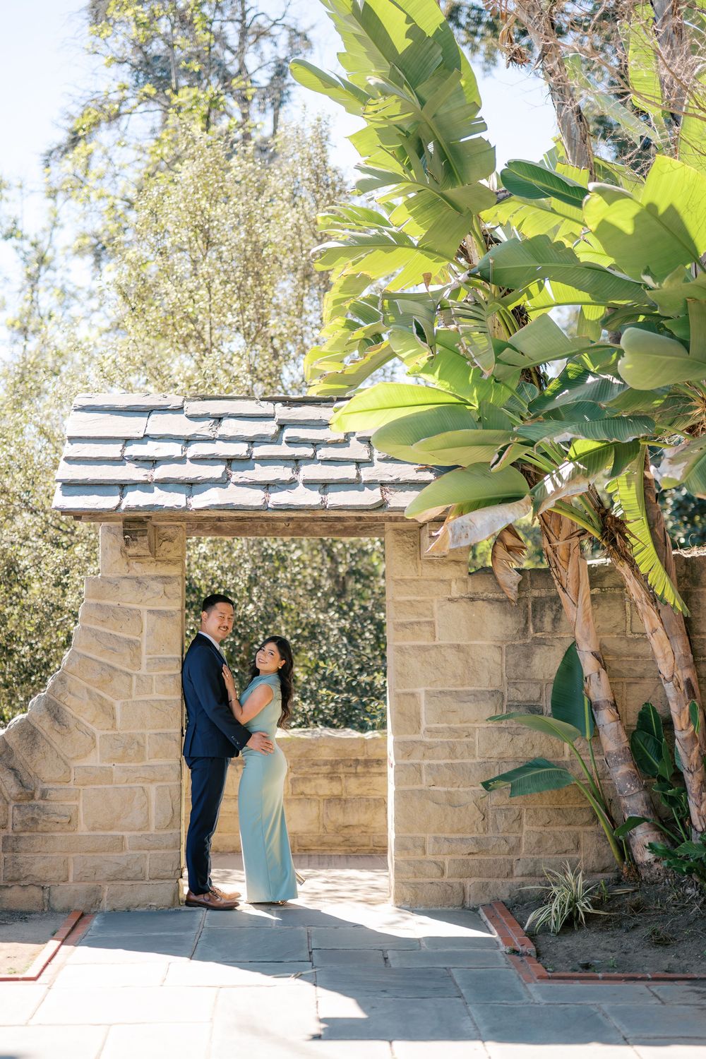 A couple shares a tender moment under a stone archway surrounded by lush greenery and sunlight.