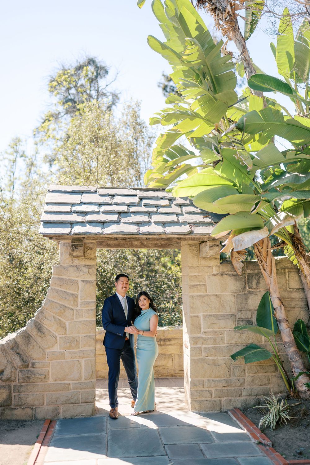A couple poses together against a stone wall backdrop with tropical plants in an outdoor garden setting.
