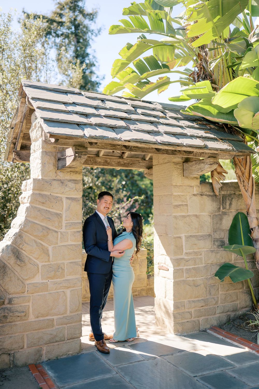 A couple in formal attire shares romantic moments against a stone wall backdrop in a garden setting.