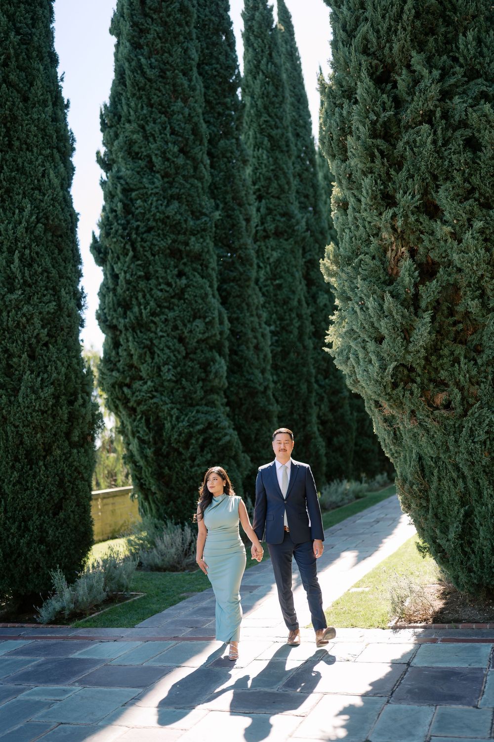 A couple walks along a cypress tree-lined path in a scenic Italian garden setting.