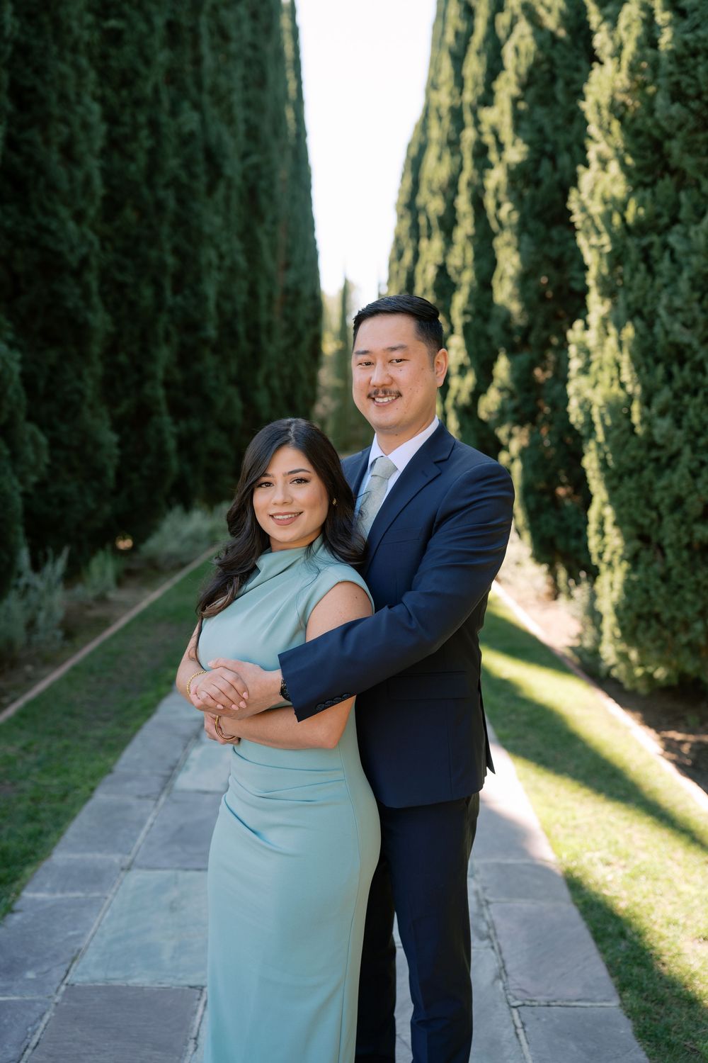 A couple embraces on a garden path lined with tall cypress trees on a sunny afternoon.