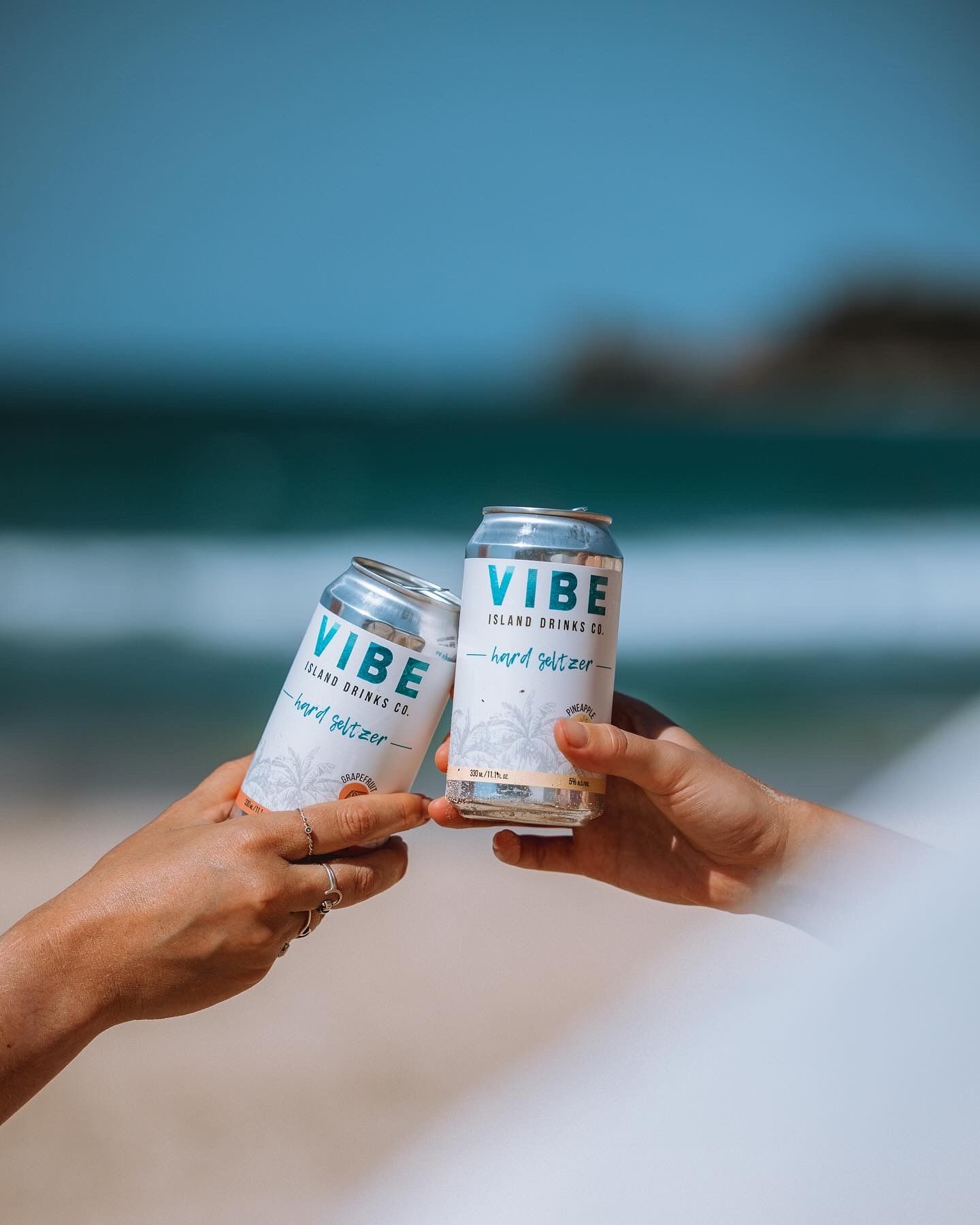 Hands toasting with cans of Vibe hard seltzer against ocean backdrop.