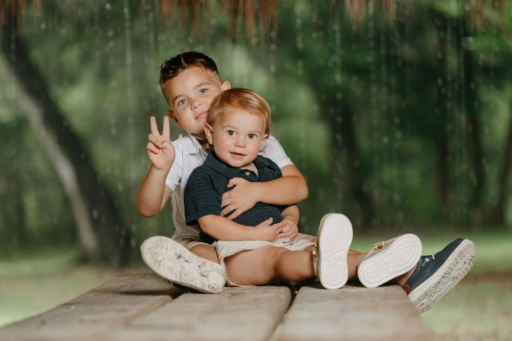 Two young brothers sitting on a wooden deck outdoors, older boy flashing peace sign while hugging toddler sibling.