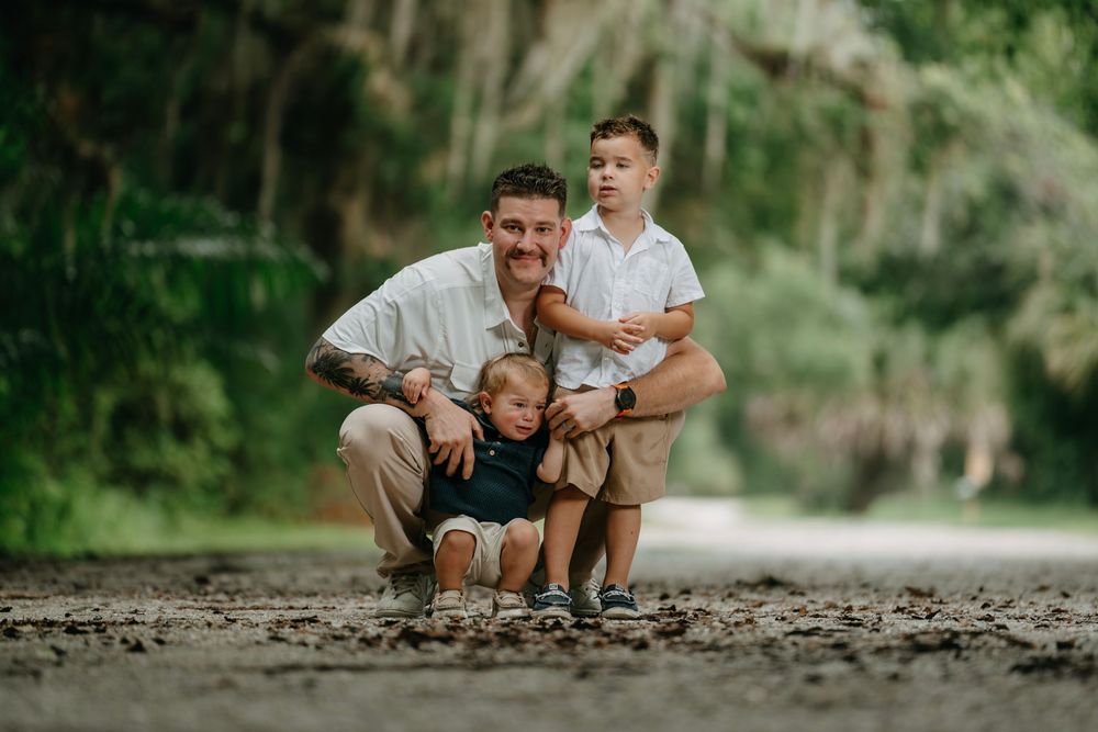 Father crouching with two young sons on a scenic outdoor path surrounded by lush green trees.