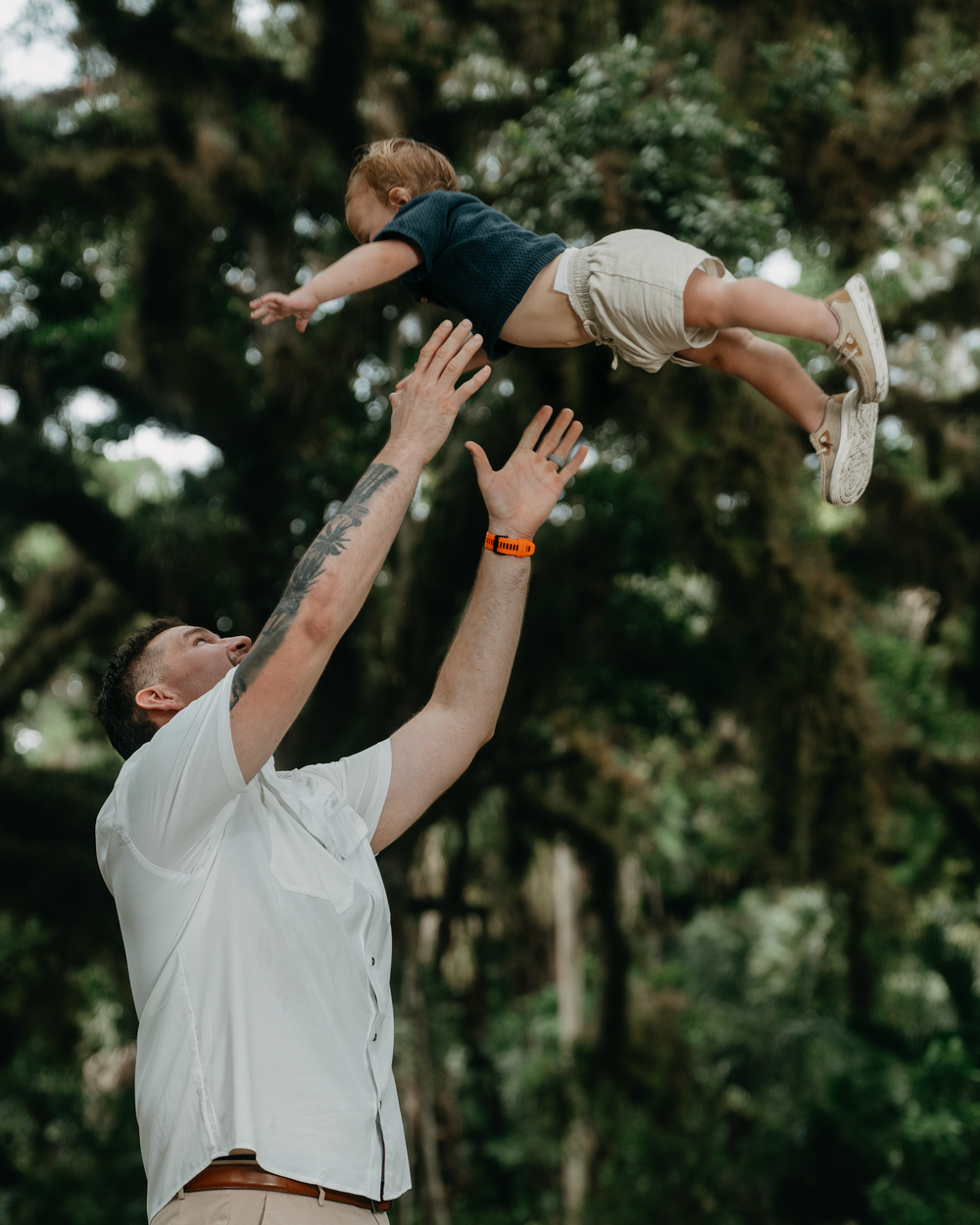 A father joyfully tosses his young child into the air outdoors, surrounded by lush green trees.