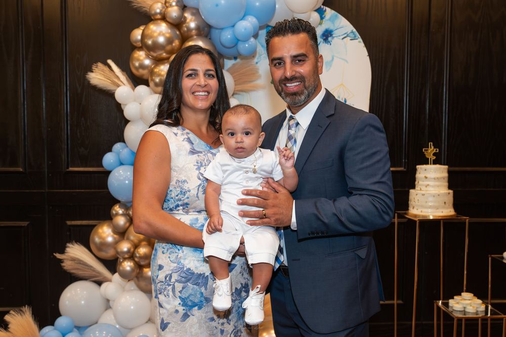 Family posing at a blue and gold balloon decorated birthday party, parents holding baby in white outfit.