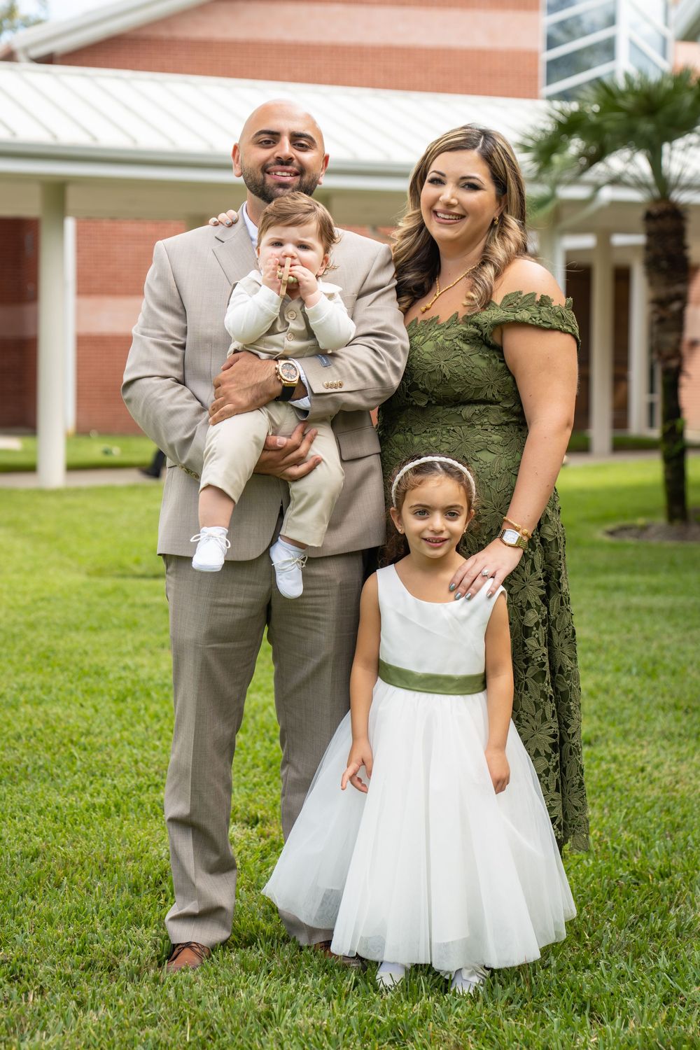 Family of four posing outdoors, parents in green and tan formal wear with two young children at a special event.