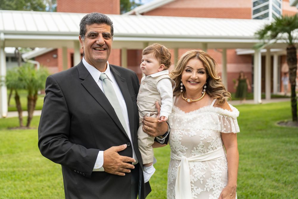 Couple in formal attire holding a baby boy outdoors at an elegant venue with lush greenery.