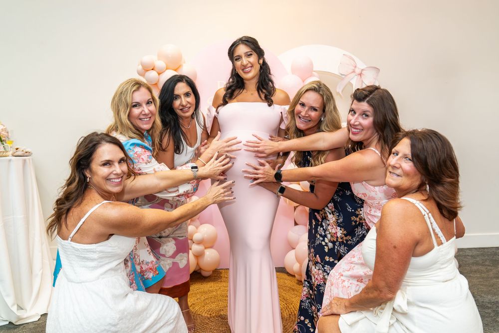 A group of women in pink and floral dresses celebrate joyfully together at a party with pink decor.