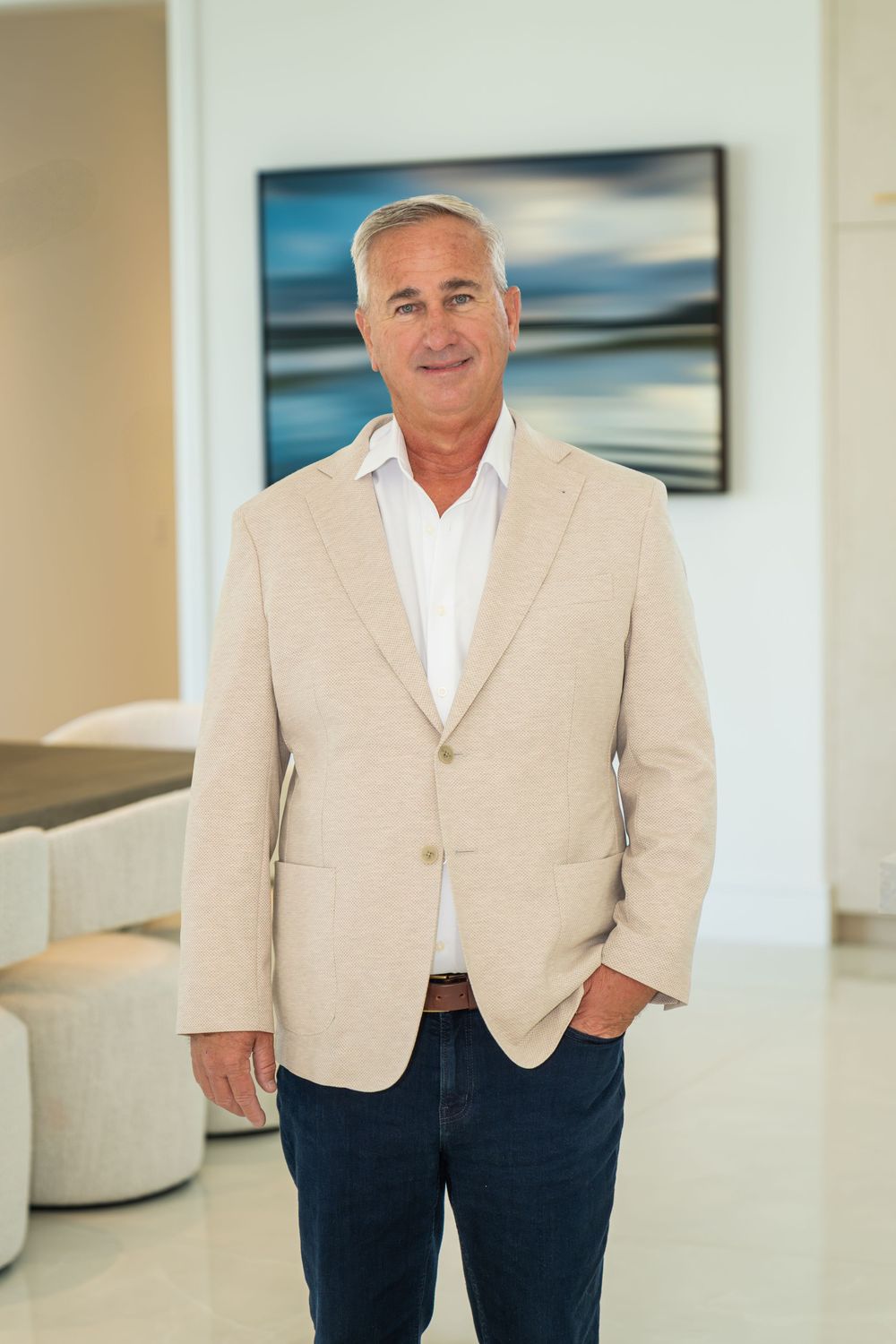 A confident middle-aged man in a beige blazer and jeans smiles while standing in a modern, bright interior space.