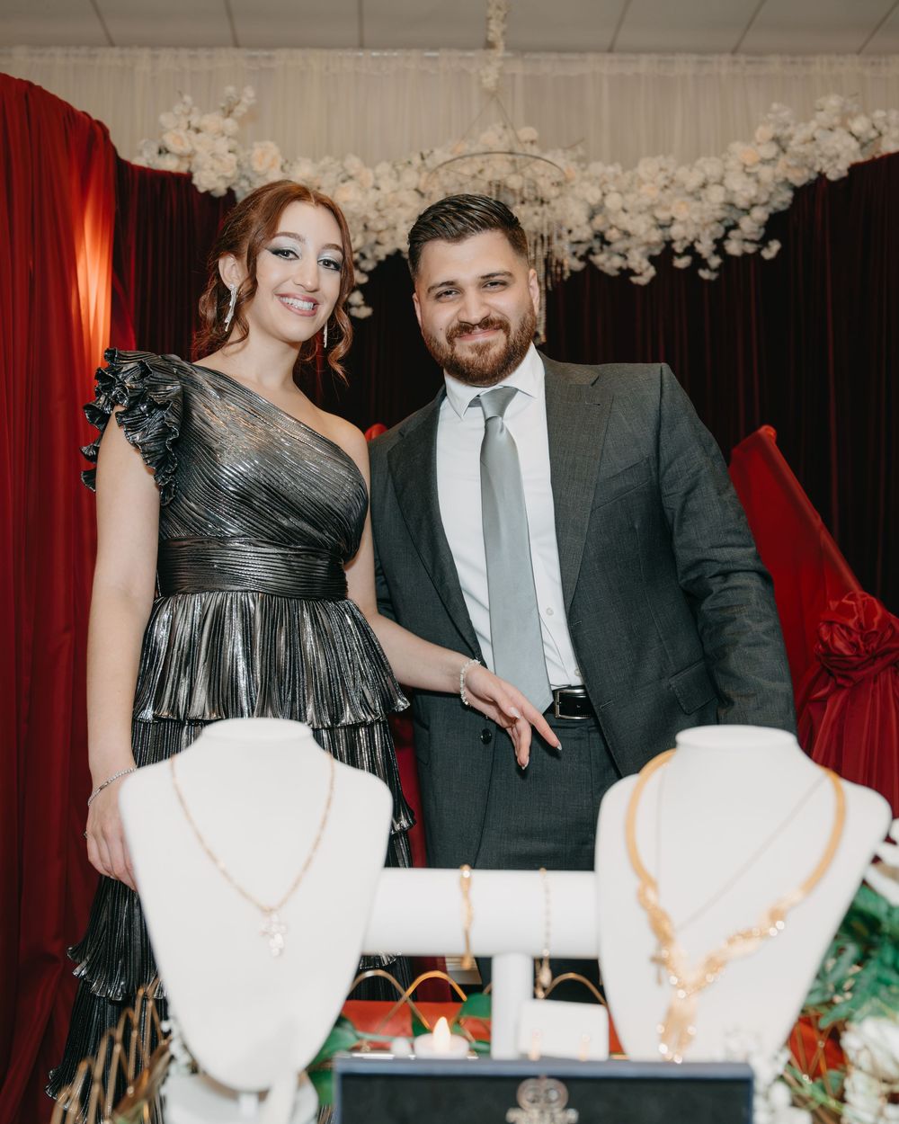 Elegant couple posing at a jewelry display with necklaces, dressed in formal attire at a decorated event venue.