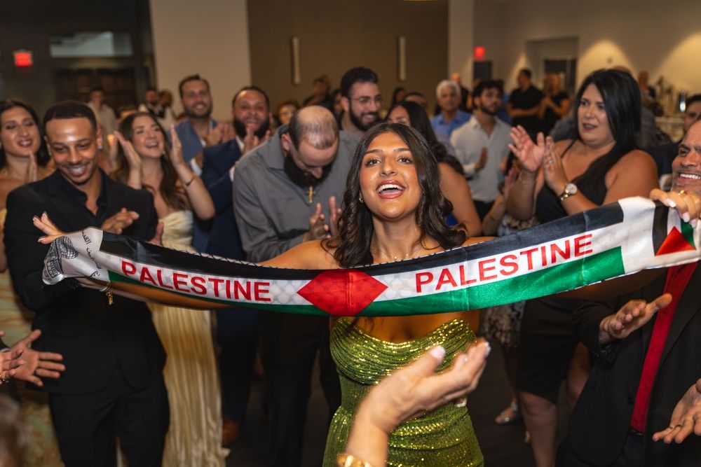 Woman in green dress holds Palestine scarf at lively celebration event with clapping crowd in background.