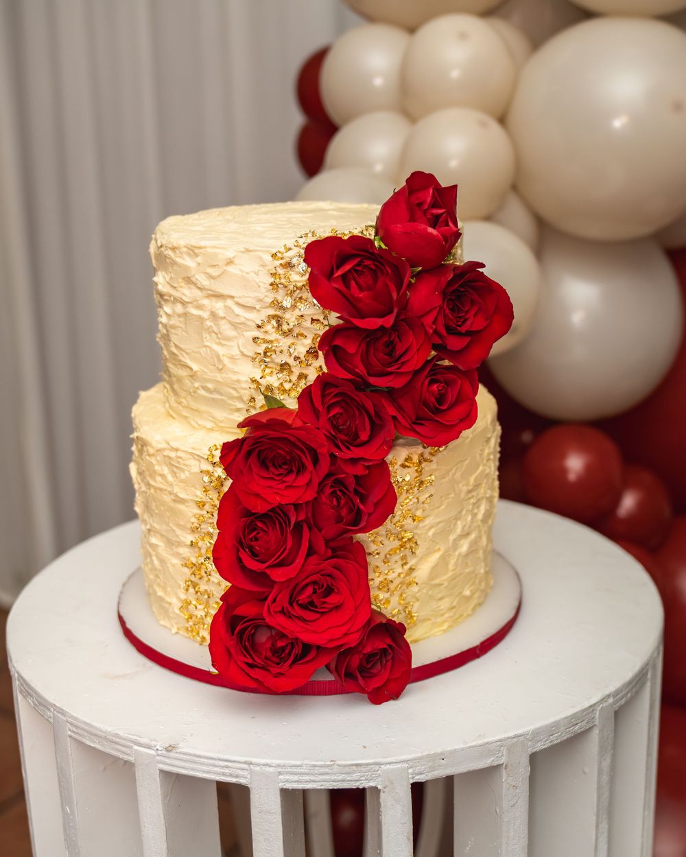 Two-tier ivory textured cake with gold leaf accents and cascading red roses on a white pedestal.