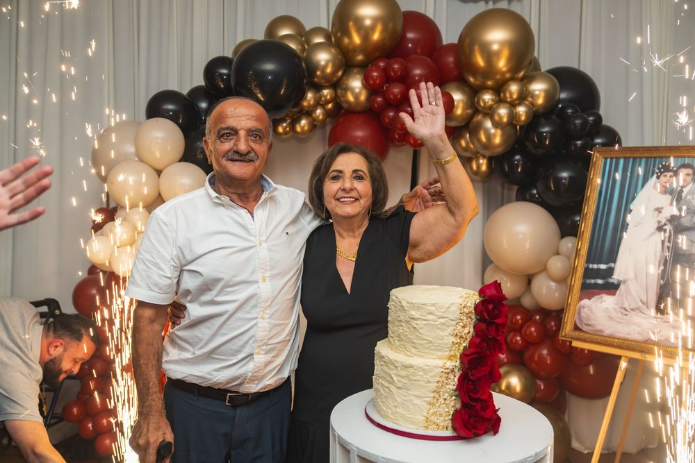 Couple smiling beside a white tiered cake with gold balloons and sparklers at an anniversary celebration.