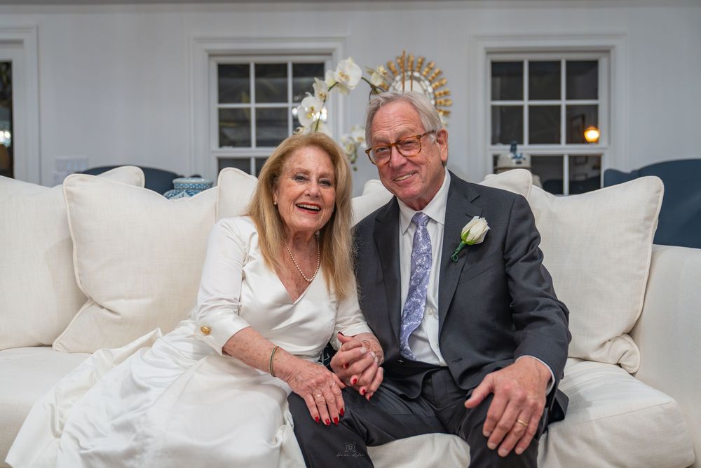 Elegant couple in formal attire sitting together on a white sofa, smiling warmly at a special celebration event.