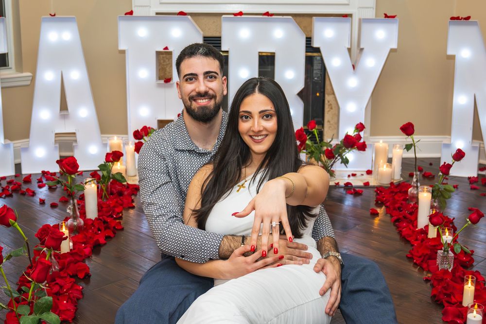 Couple poses in front of illuminated MARRY ME letters surrounded by red roses and candles after romantic proposal.