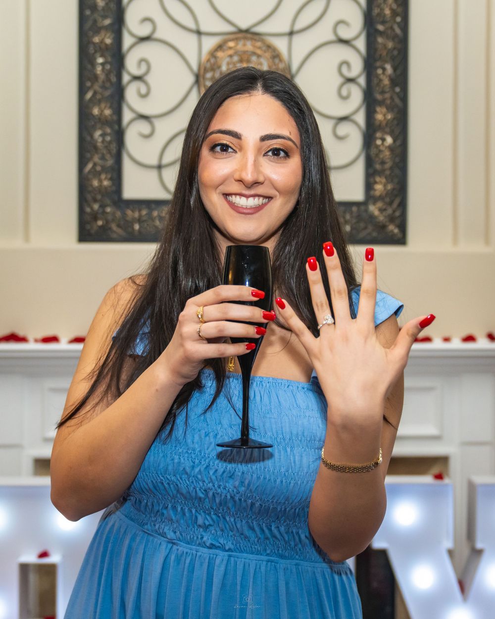 Smiling woman in blue dress showing engagement ring while holding champagne glass with red nails at romantic celebration.