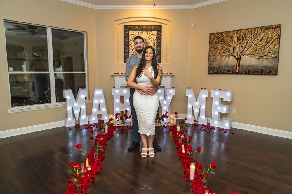 Couple poses in front of illuminated MARRY ME letters surrounded by red roses and candles in a romantic proposal setup.