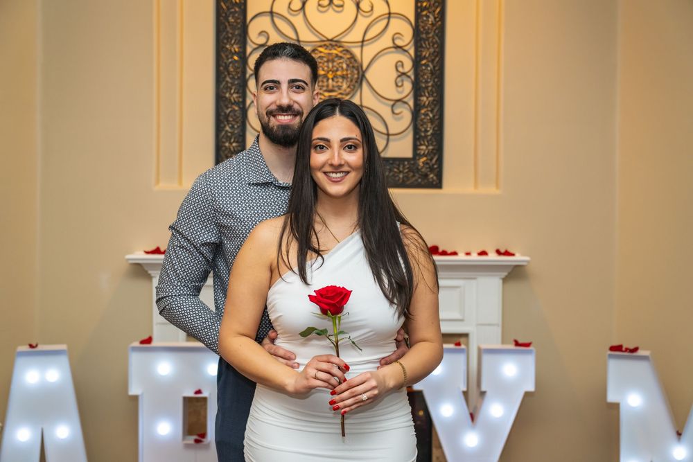 Couple posing with a red rose in front of illuminated marquee letters and a white fireplace during a romantic proposal.