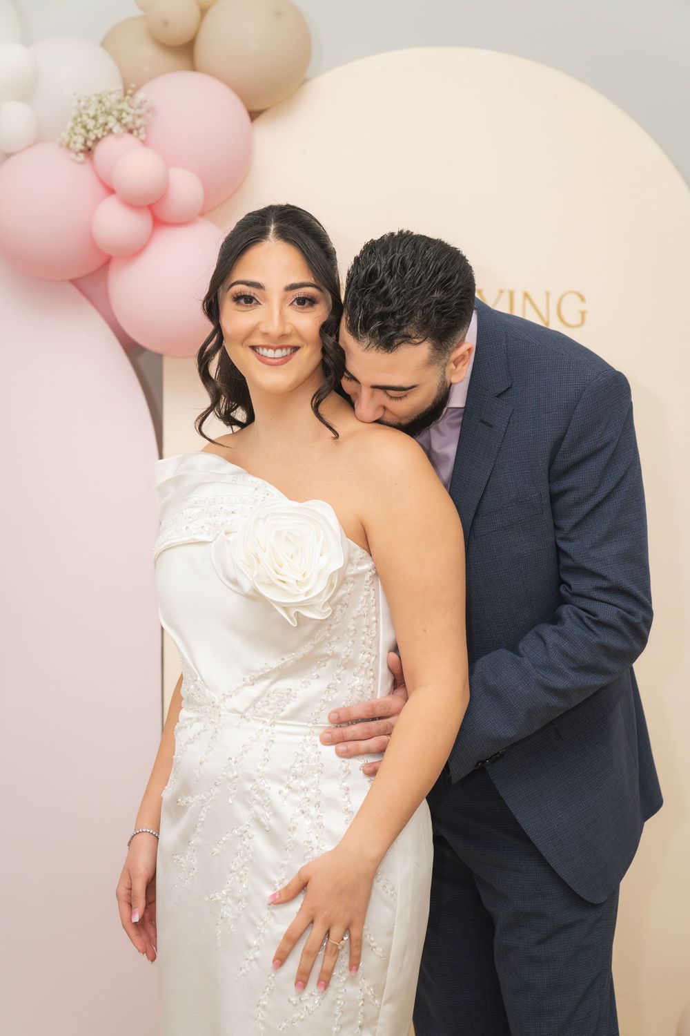 A couple in wedding attire share intimate moments against a backdrop decorated with pink and neutral balloons.