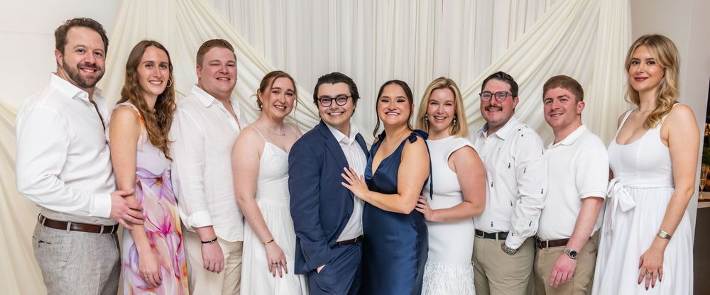 A group of elegantly dressed wedding guests posing together, smiling, in white and pastel outfits at a formal celebration.