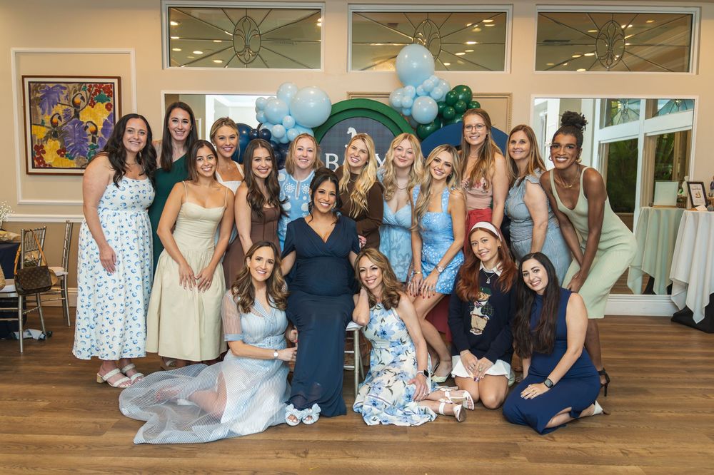 Group of women posing together at a baby shower with blue balloon arch decor in elegant venue.