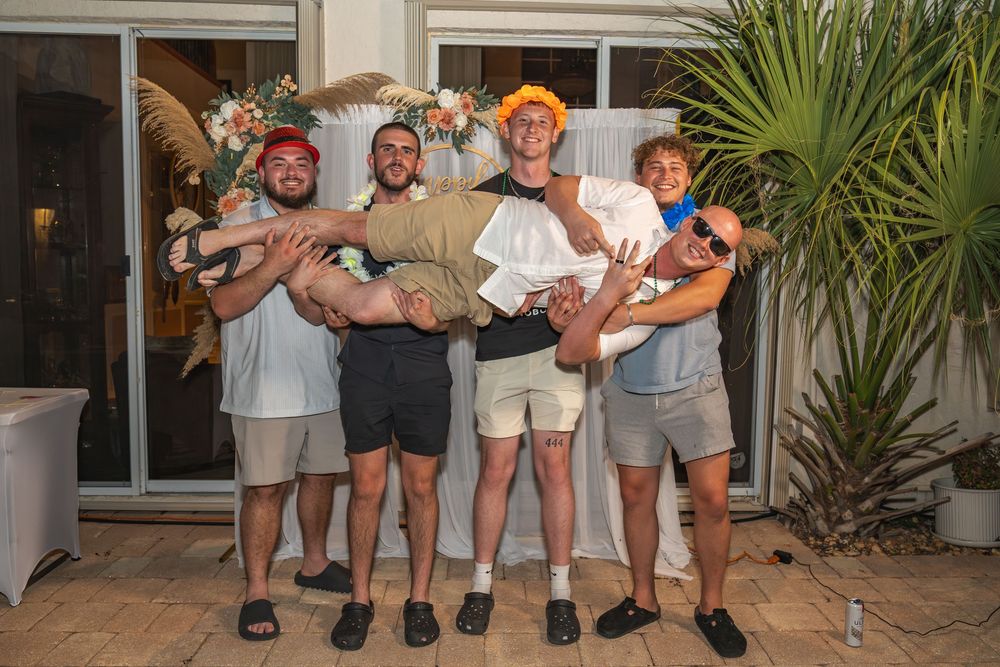 Group of young men posing outdoors at a party, one being lifted, wearing casual shorts and colorful hats.