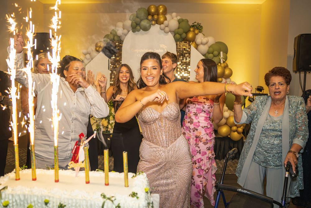 Two women in elegant dresses celebrate a birthday party with sparklers and a large cake at a festive event.