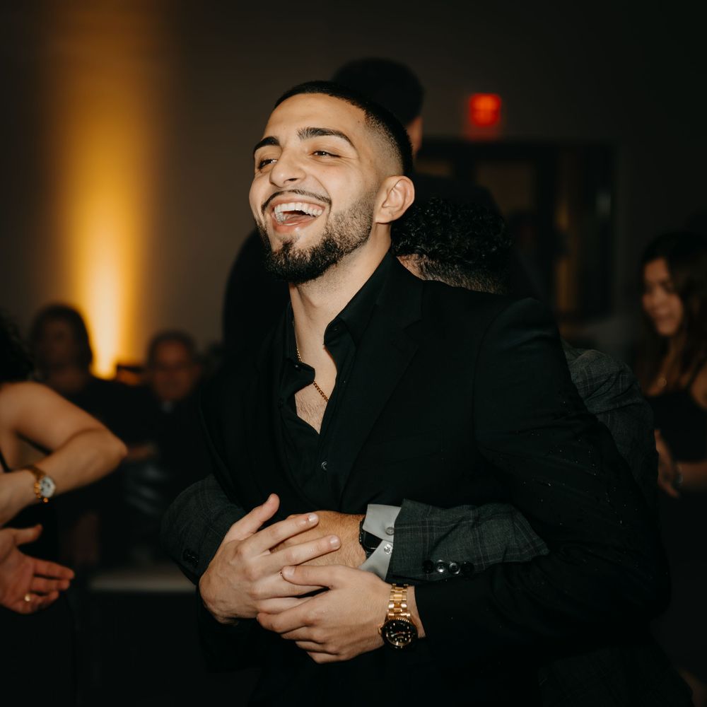 A smiling young man with a beard wearing a black shirt and gold watch laughs joyfully at an indoor event.