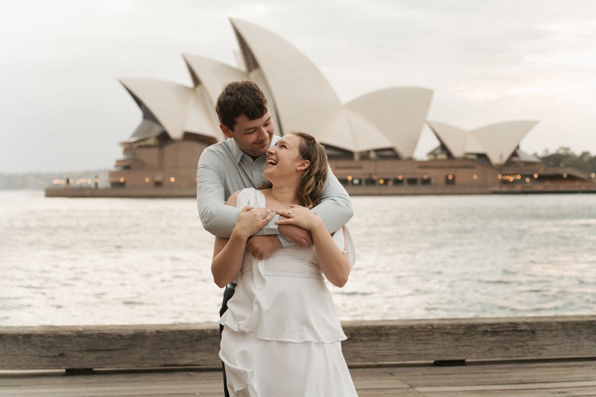 A romantic couple embraces in front of the iconic Sydney Opera House during sunset.