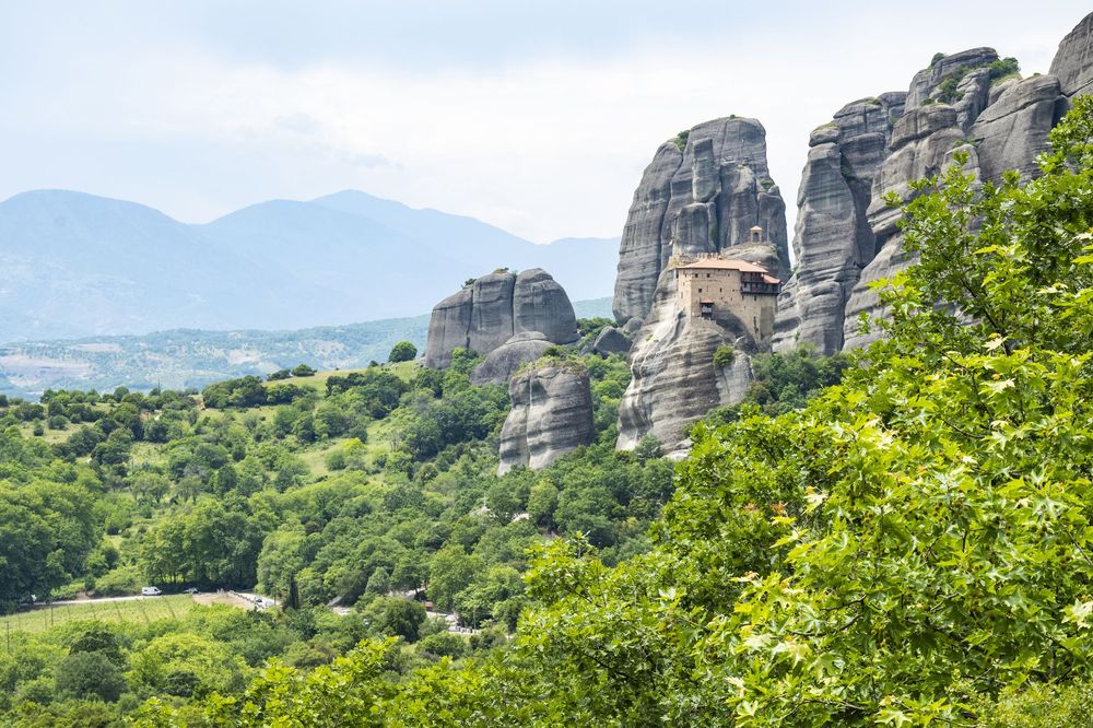 Dramatic rock formations rise above green forest landscape at Meteora in Greece with misty mountains in background.